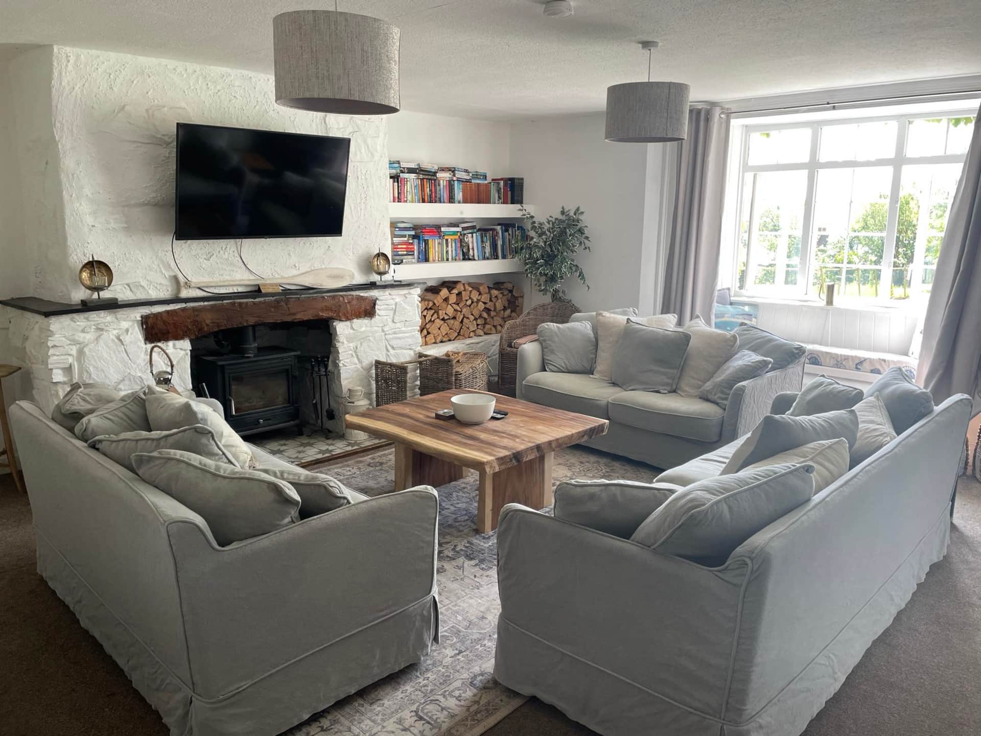 The Farmhouse Fowey Living room with log burner, beautifully furnished with grey sofas, a rustic wooden coffee table, and a TV on a brick wall. Views out of he garden from the window