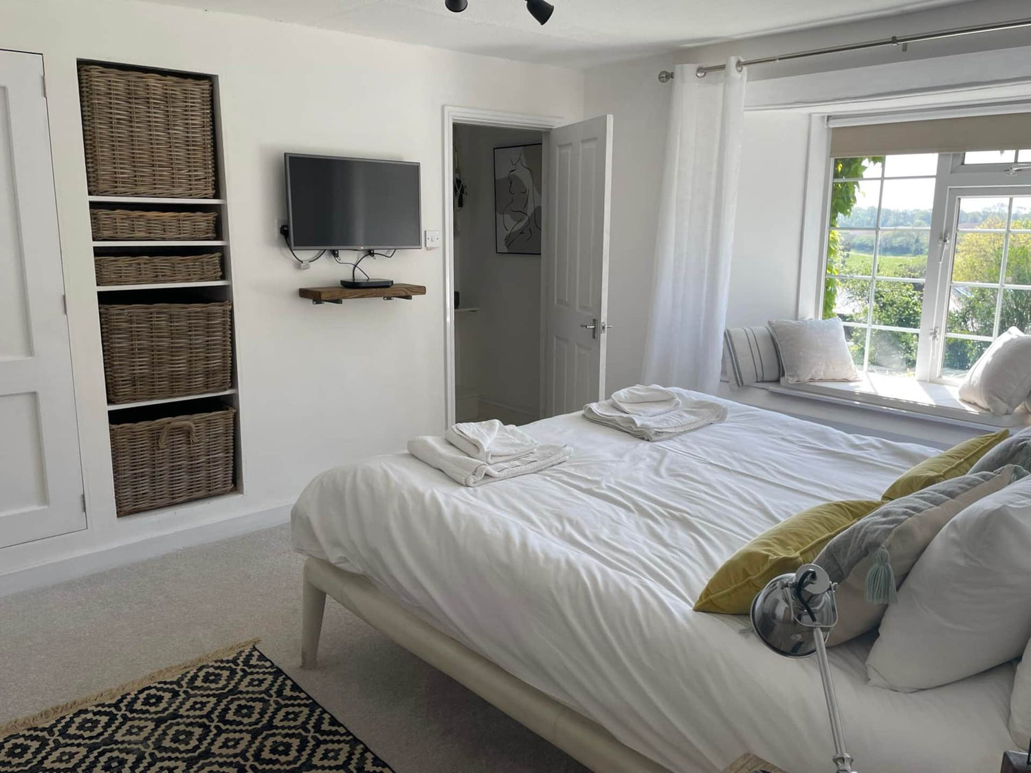 The Farmhouse Primary Bedroom with countryside view. Image shows the double bed, TV, and wicker storage baskets and door to en-suite bathroom. 