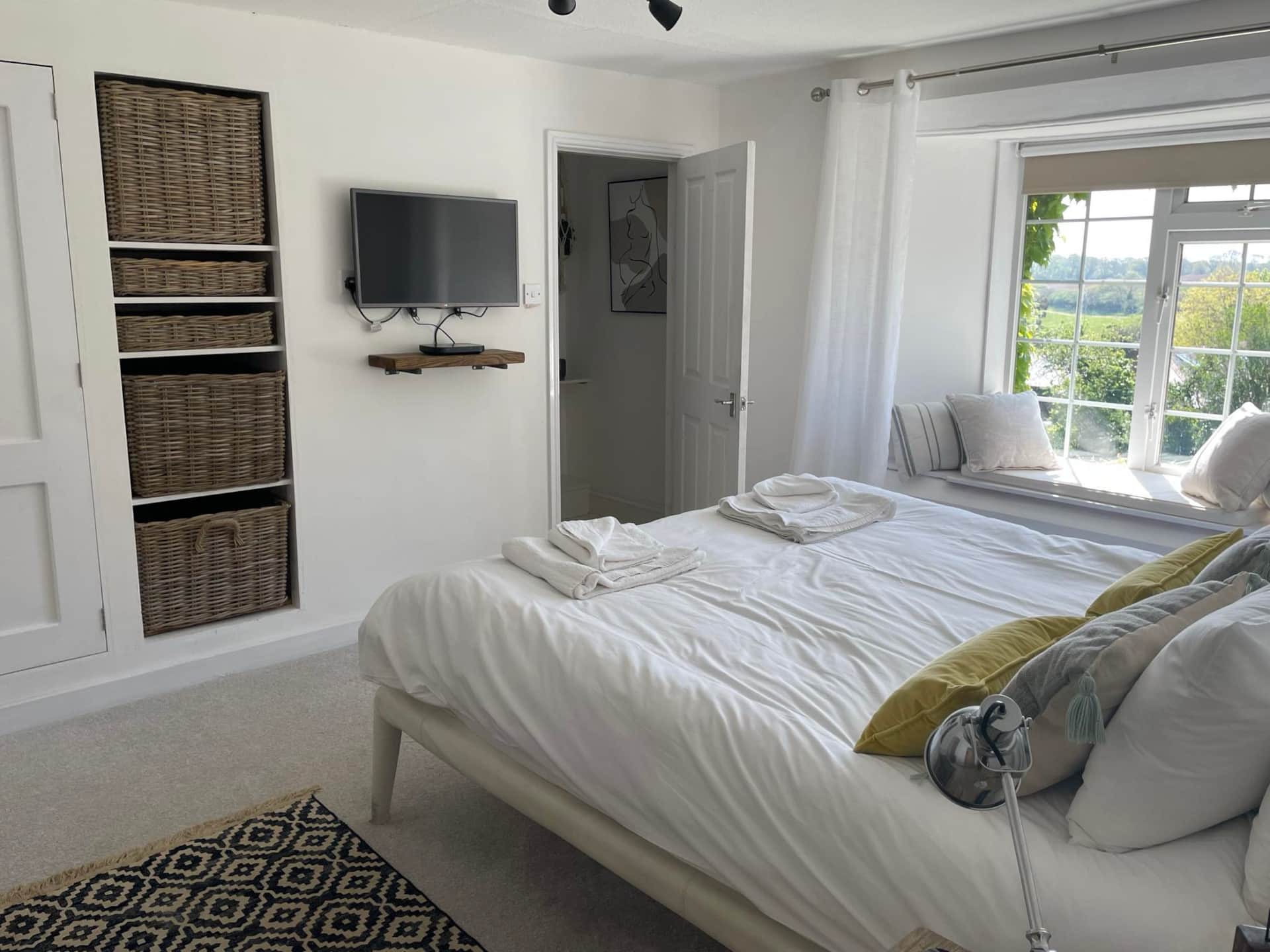The Farmhouse Primary Bedroom with countryside view. Image shows the double bed, TV, and wicker storage baskets and door to en-suite bathroom. 